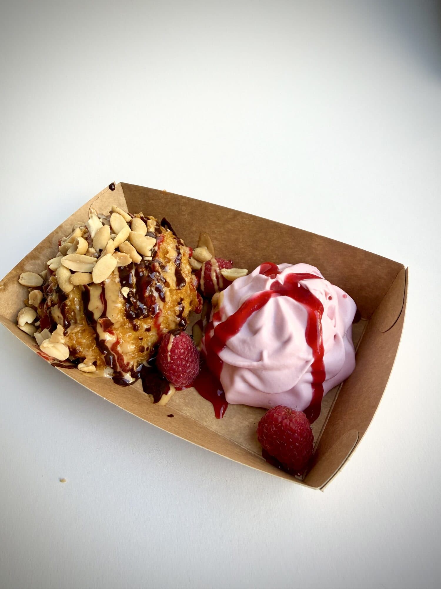 A fried scoop of banana ice cream with roasted peanuts and chocolate sauce, next to a pink raspberry-lime cream with raspberry sauce and fresh raspberries, in a kraft paper tray.