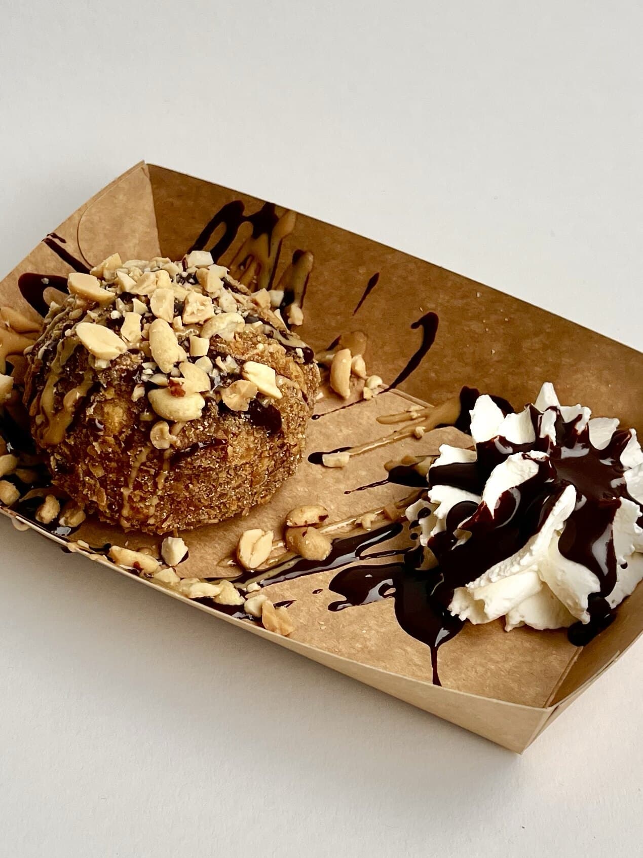 Pallina di gelato alla banana fritta con arachidi tostate e salsa al cioccolato, accanto a un ciuffo di panna con salsa al cioccolato, in una vaschetta di carta kraft.