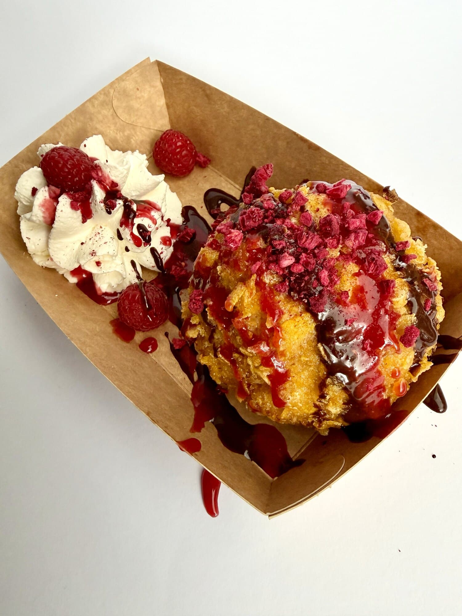 Pallina di gelato alla vaniglia fritta con salsa al lampone e polvere di lampone, accanto a un ciuffo di panna con lamponi freschi, in una vaschetta di carta kraft.